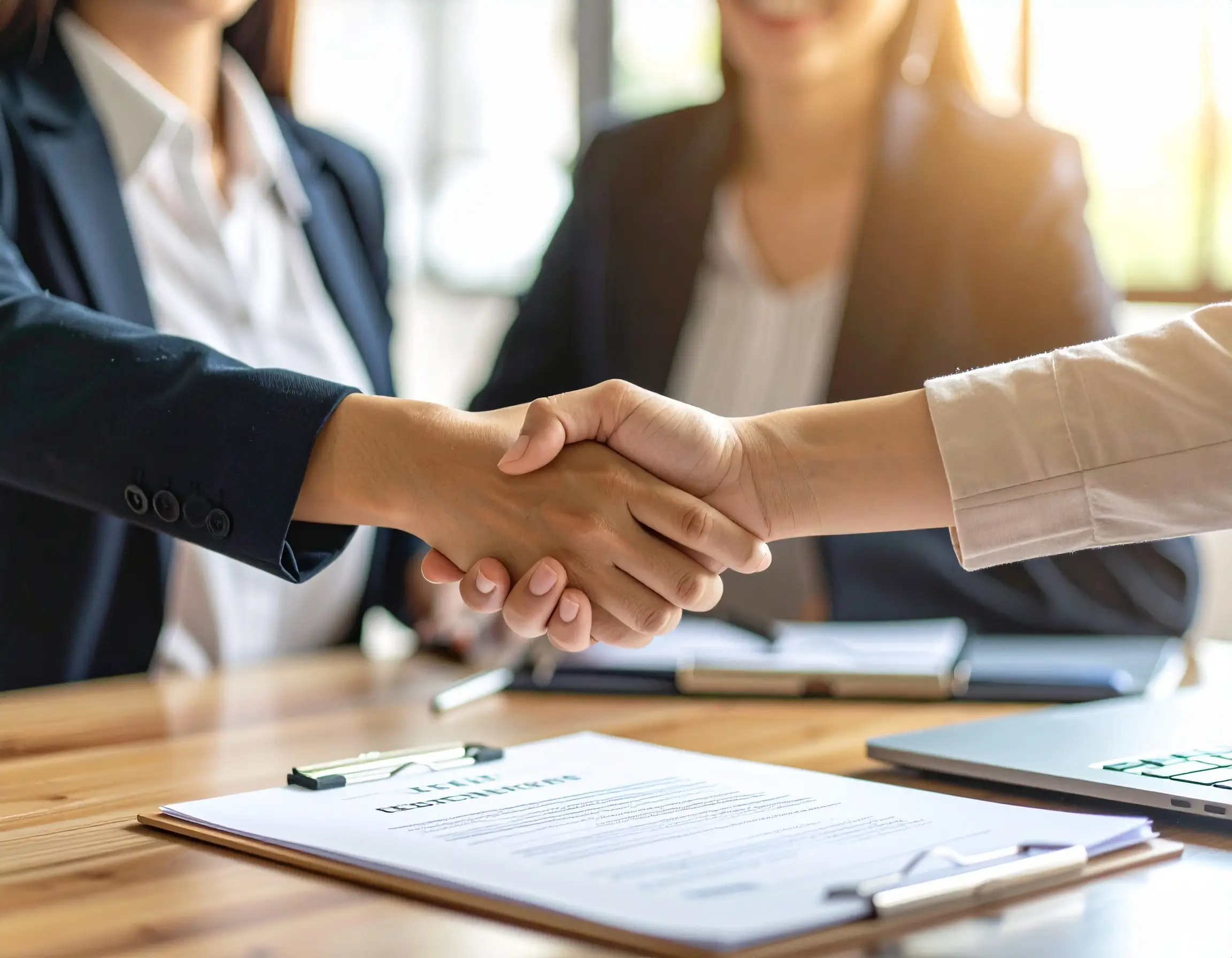 Two individuals shaking hands over a table with a signed agreement and a laptop nearby, suggesting a professional agreement related to the first time home buying process.